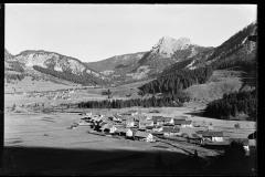 Ortschaft umgeben von hohen, bewaldeten Bergen und Grünfläche.