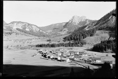 Ortschaft umgeben von hohen, bewaldeten Bergen und Grünfläche.
