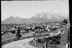 Ortschaft umgeben von hohen, bewaldeten Bergen und Grünfläche.