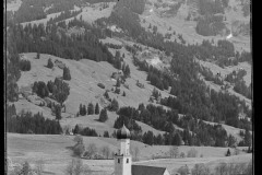 Ortschaft mit Kirche umgeben von hohen, bewaldeten Bergen und Grünfläche.