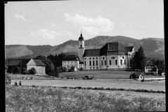 Ortschaft mit Kirche umgeben von hohen, bewaldeten Bergen und Grünfläche.