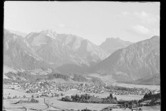 Große Ortschaft umgeben von hohen, bewaldeten Bergen und Grünfläche.