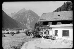 Großes Haus mit Terrasse mit Tischen und Stühlen, dahinter eine Ortschaft mit Kirche und Bergblick
