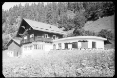 Großes Haus mit Terrasse mit Tischen und Stühlen, daneben ein Holzstall und der Wald