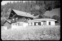 Großes Haus mit Terrasse mit Tischen und Stühlen, daneben ein Holzstall und der Wald