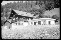 Großes Haus mit Terrasse mit Tischen und Stühlen, daneben ein Holzstall und der Wald