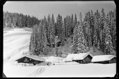 Dick verschneite Holzhütten, dahinter Menschen und Skispuren im Schnee
