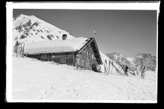 Hölzerne Skihütte, mit viel Skiequipment und Menschen vor den Dreischafsalpköpfen