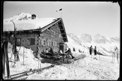 Hölzerne Skihütte, voll besetzte Terasse und Tische mit Menschen, viel Skiequipment, vor den Dreischafsalpköpfen