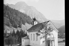 Kapelle mit Turm, auf einer Wiese, dahinter die Berge