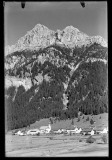 Ortszentrum mit Kirche und Wohnhäusern, auf einem Feld, von Bergen umgeben