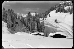 Dick verschneite Holzhütten, dahinter Menschen und Skispuren im Schnee
