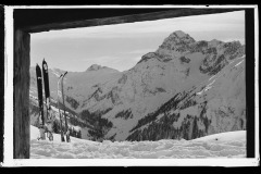 Verschneite Almhütte, umgeben von Bergen und Wald, inmitten von Skispuren durchzogenen Feld