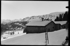 Gekacheltes und Holzverkleidetes Haus umgeben von Schneefeld, mit Skispuren durchzogen, dahinter der Wald