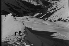 Zwei Erwachsene mit Ski Ausrüstung bepackt, am Rande einer Skiabfahrt die ins bewaldete Tal führt, dahinter die Berge