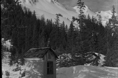 Aus Stein gemeißelter Bildstock mit Fenster und gekachelt dem Dach, davor Ski Equipment aus Holz, dahinter der Wald und eine Bergkulisse, Himmel