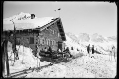 Hölzerne Skihütte, voll besetzte Terasse und Tische mit Menschen, viel Skiequipment, vor den Dreischafsalpköpfen
