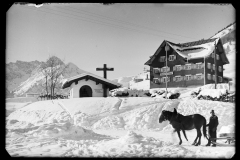 Kleine Kapelle in Hirschegg mit Holzkreuz und einem Pferd, das einen Schlitten zieht, daneben ein Haus und Berge